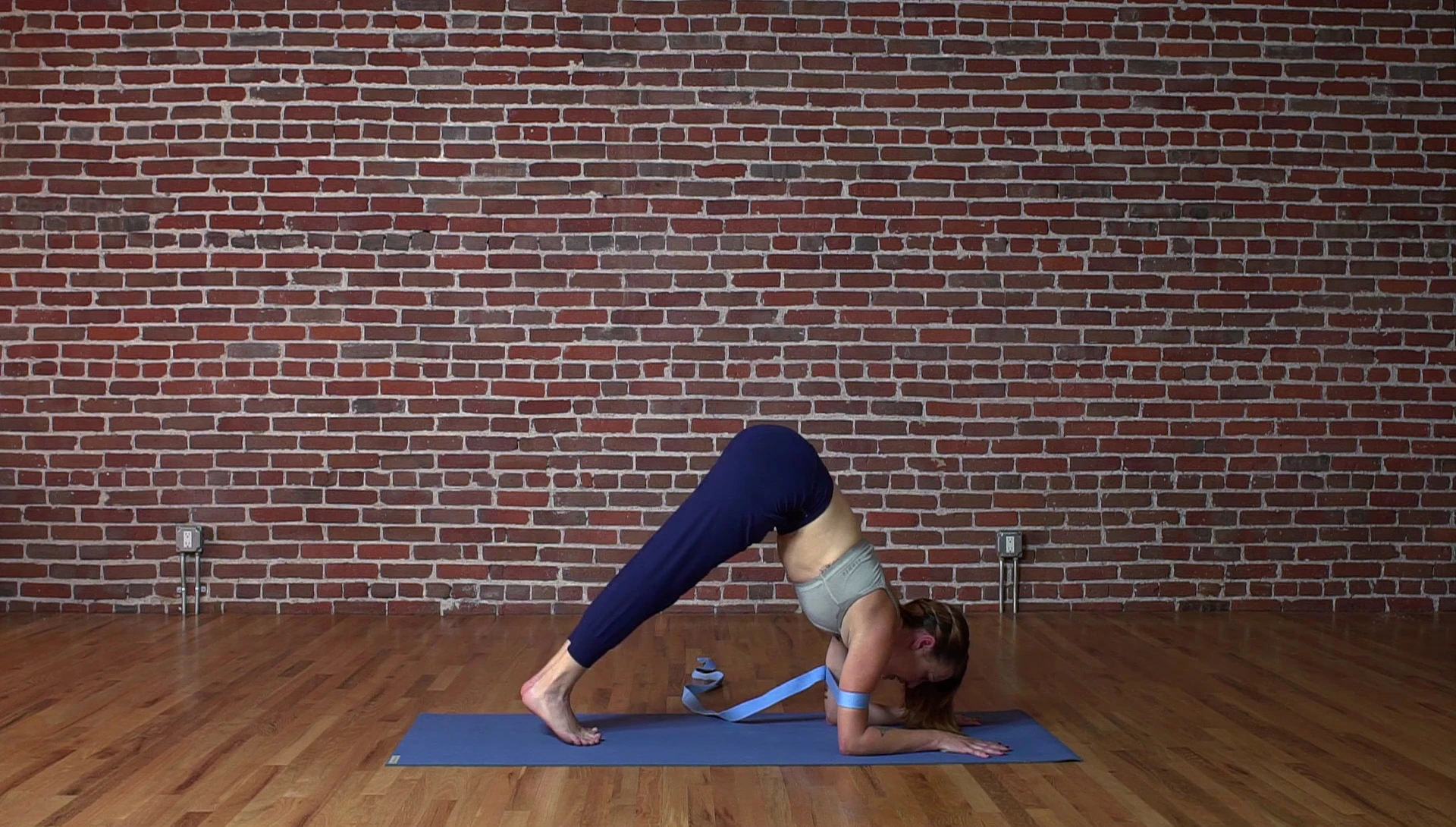 Woman lifting one leg in forearm stand prep with yoga strap.