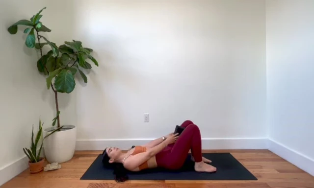 Woman lying on her back with a yoga block between her inner thighs, preparing for a glute bridge.