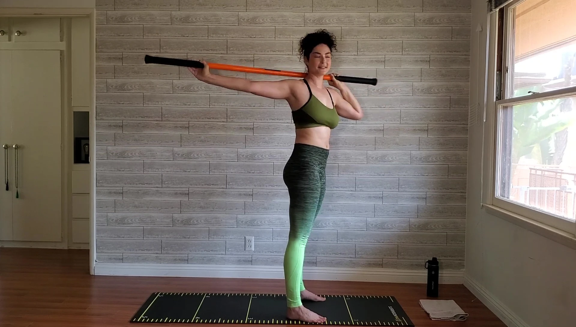 Woman performing hip and leg stretches with the stick behind her head.